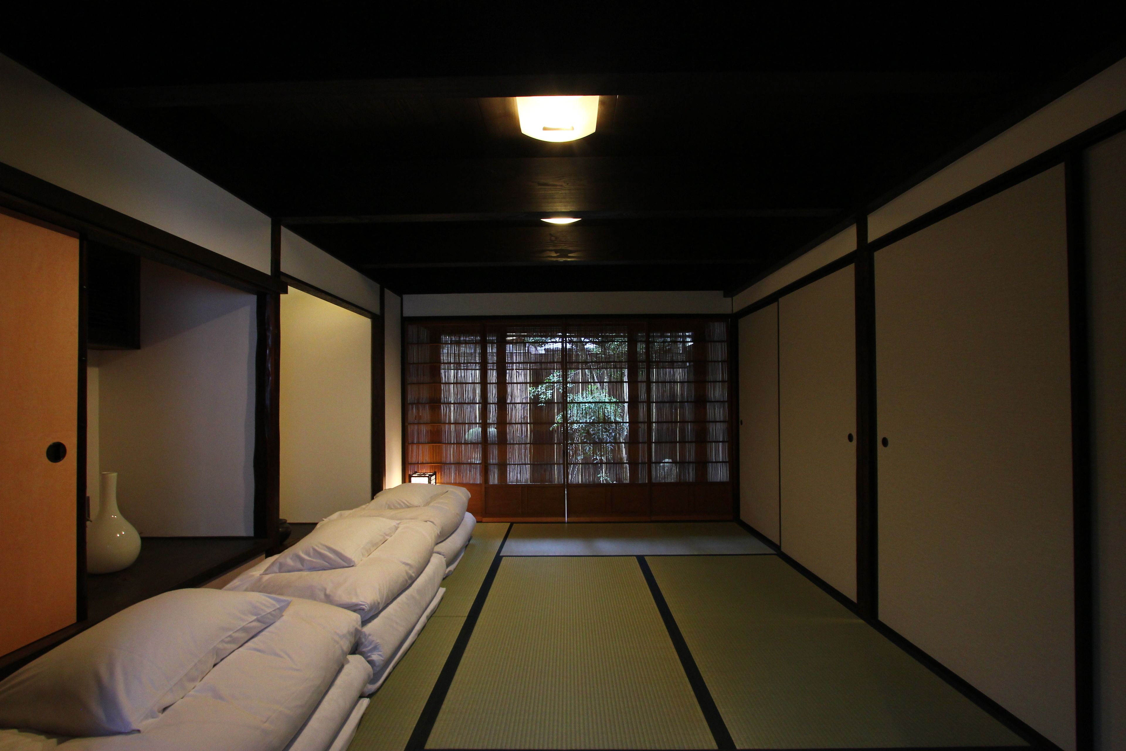 Une pièce de style japonais traditionnel, avec un sol recouvert de tatamis, une petite table sur laquelle est posé un vase, et des portes coulissantes donnant sur un jardin.