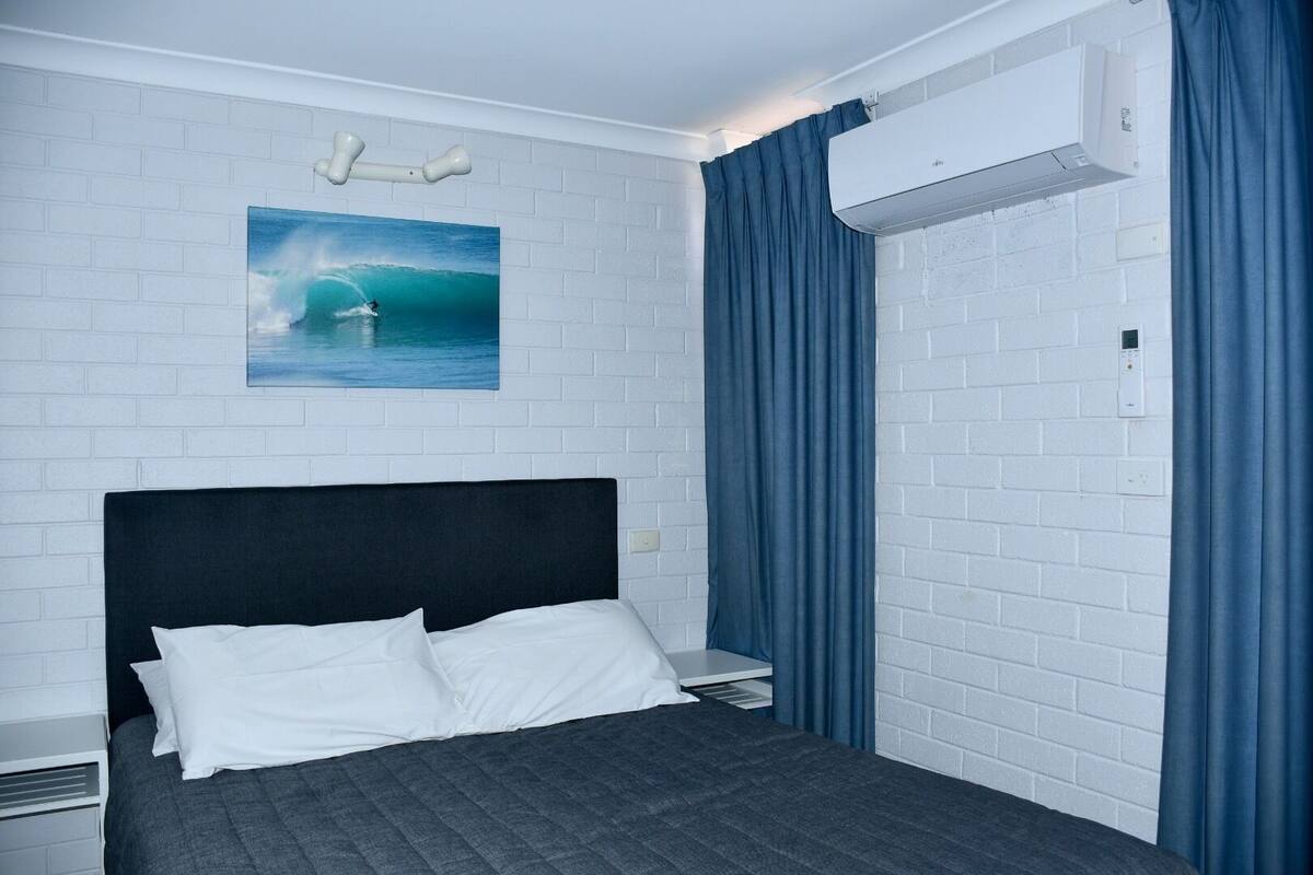 Habitación de hotel con una cama, una pared de ladrillo blanco, un cuadro de un surfista y un equipo de aire acondicionado.