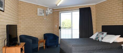 A hotel room with a brick wall, a large bed with grey bedding, a blue armchair, a wooden side table, a television, and a ceiling fan.