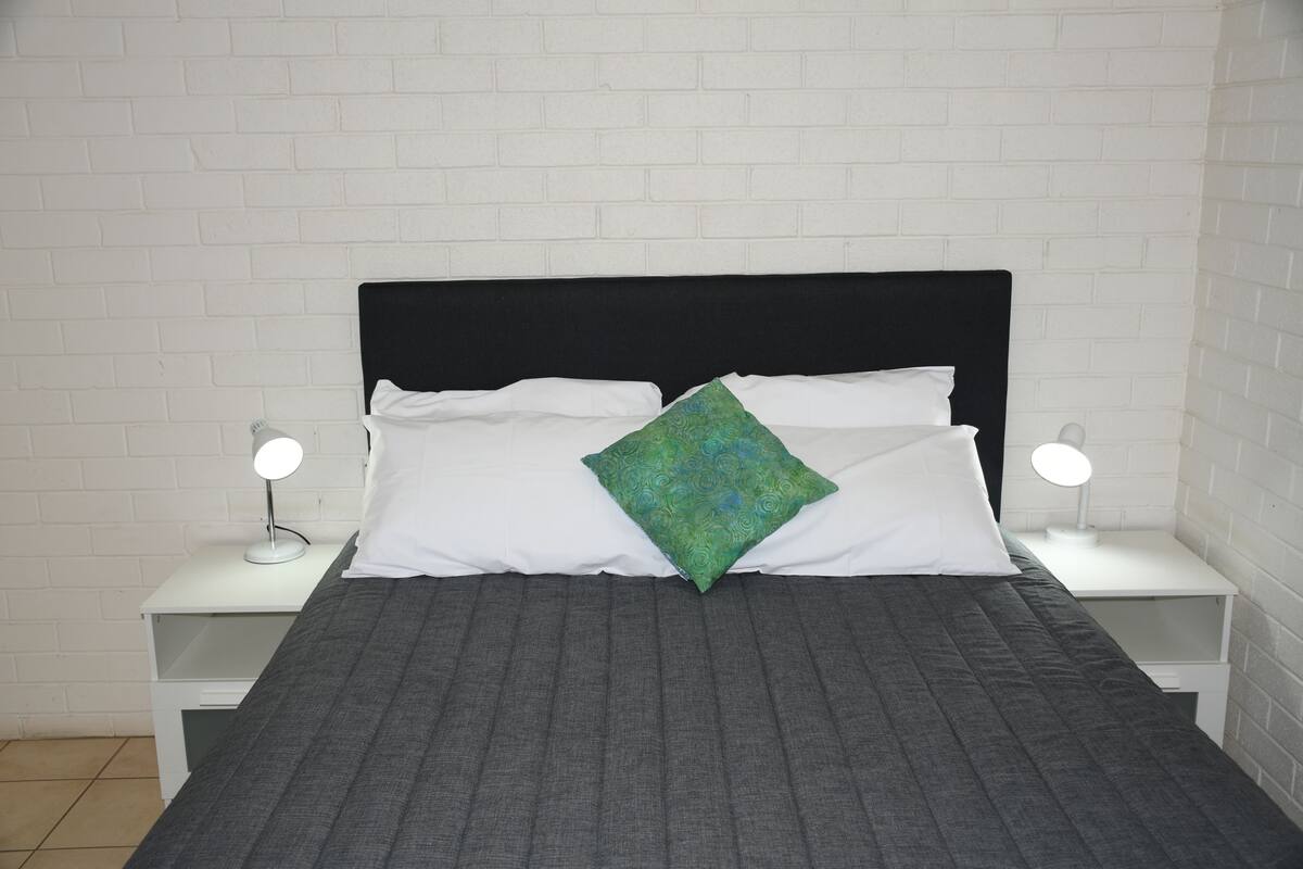 Una cama con cabecera negra, almohadas blancas y una almohada verde estampada, además de dos mesitas de noche con lámparas.