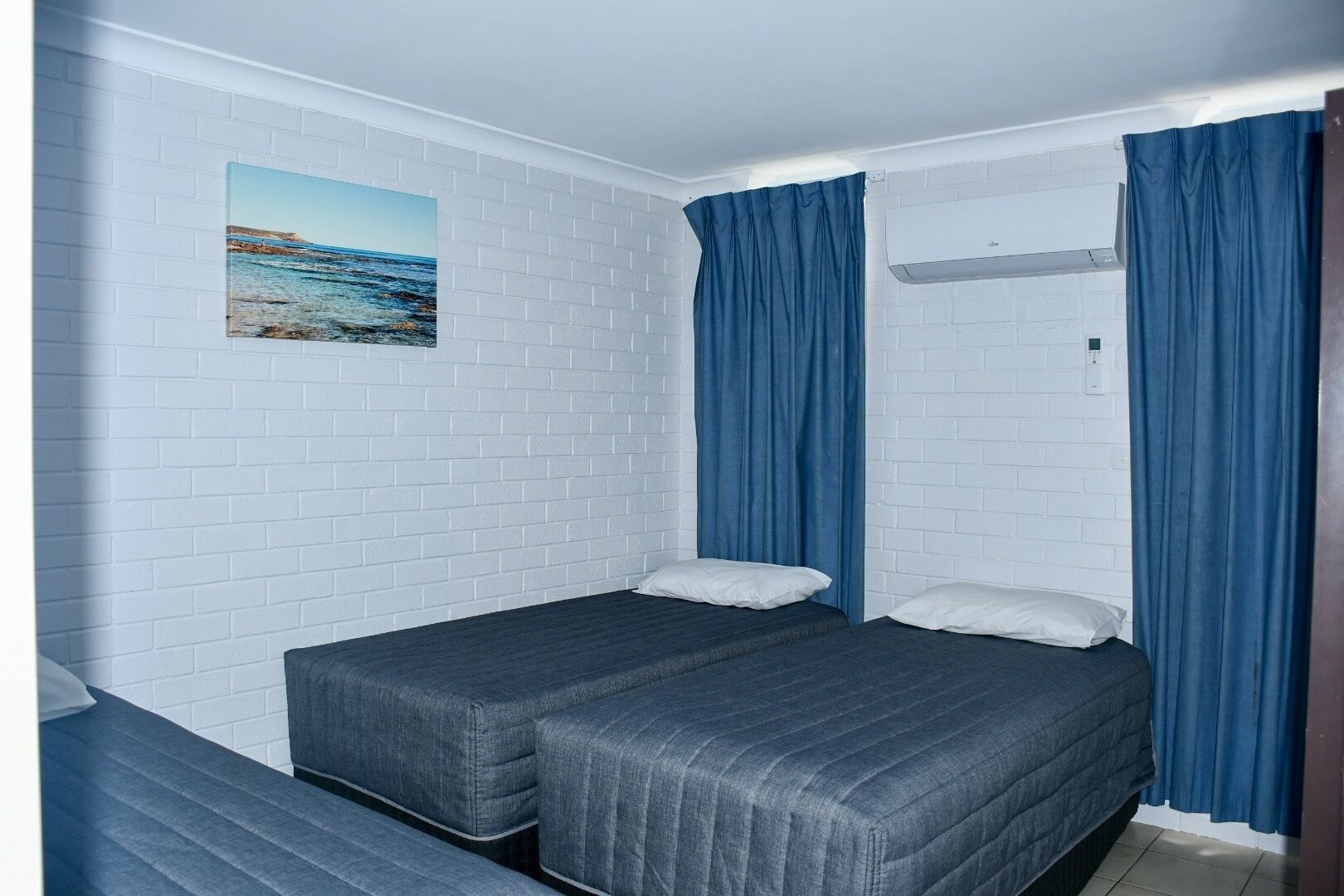 A small, clean room with white brick walls, two single beds with dark bedding, and a painting of a coastal scene.