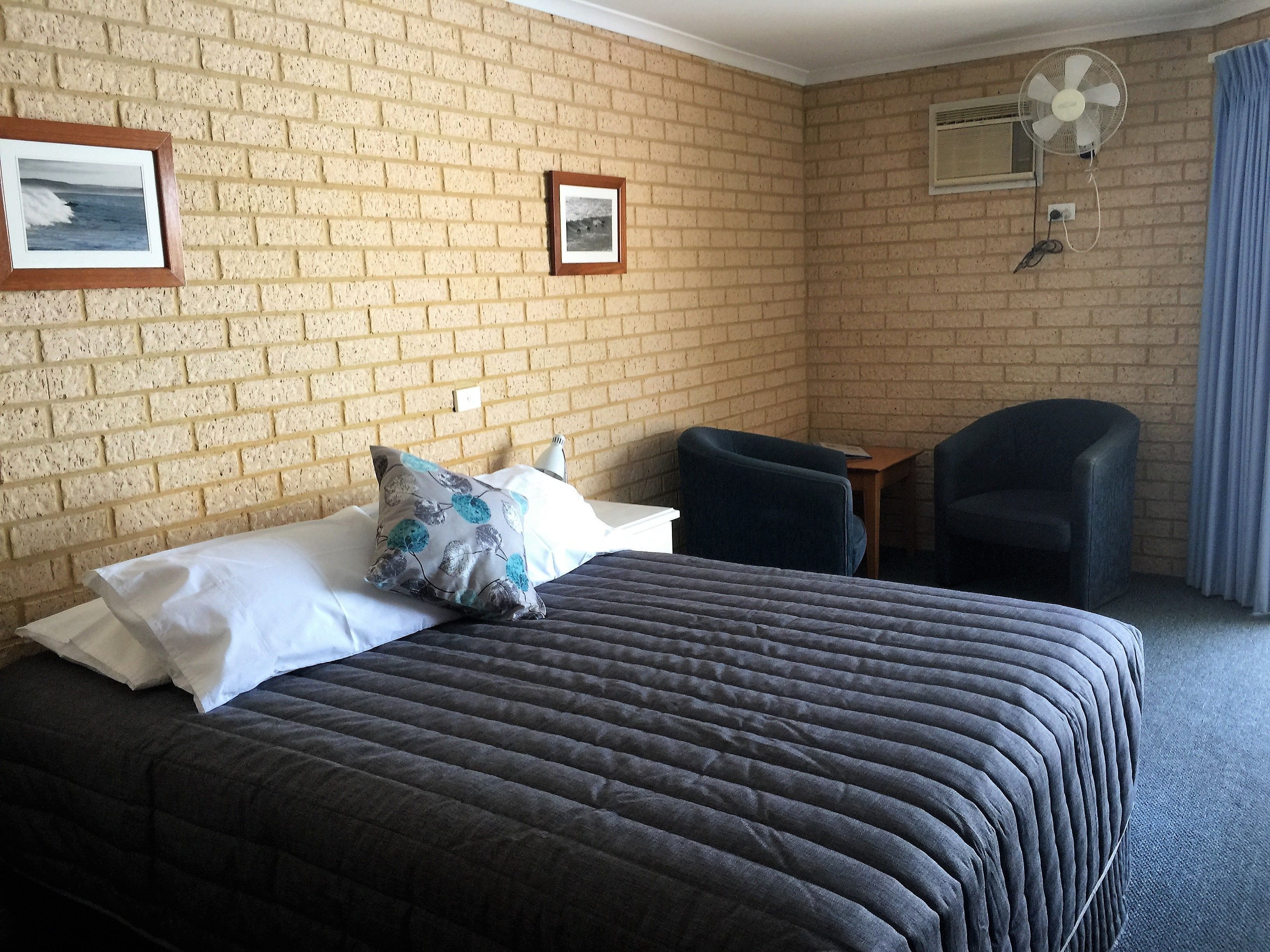 Una habitación de hotel con pared de ladrillo, una cama con colcha oscura, dos sillas, una mesita y un ventilador de techo.