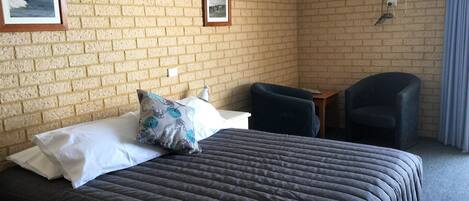 Una habitación de hotel con pared de ladrillo, una cama con colcha oscura, dos sillas, una mesita y un ventilador de techo.