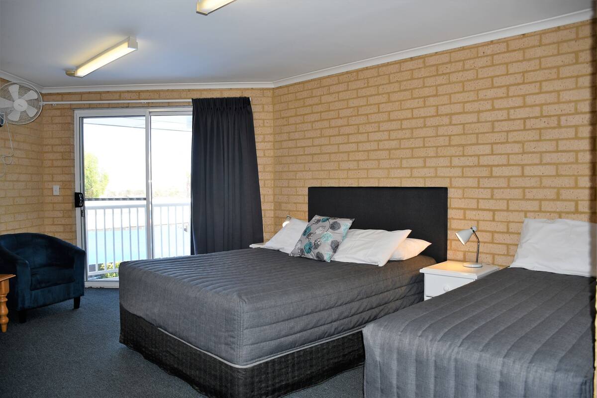 Habitación de hotel con pared de ladrillo, cama grande con ropa de cama gris, mesita de noche con lámpara, una silla y puerta corrediza de vidrio que da a un balcón.