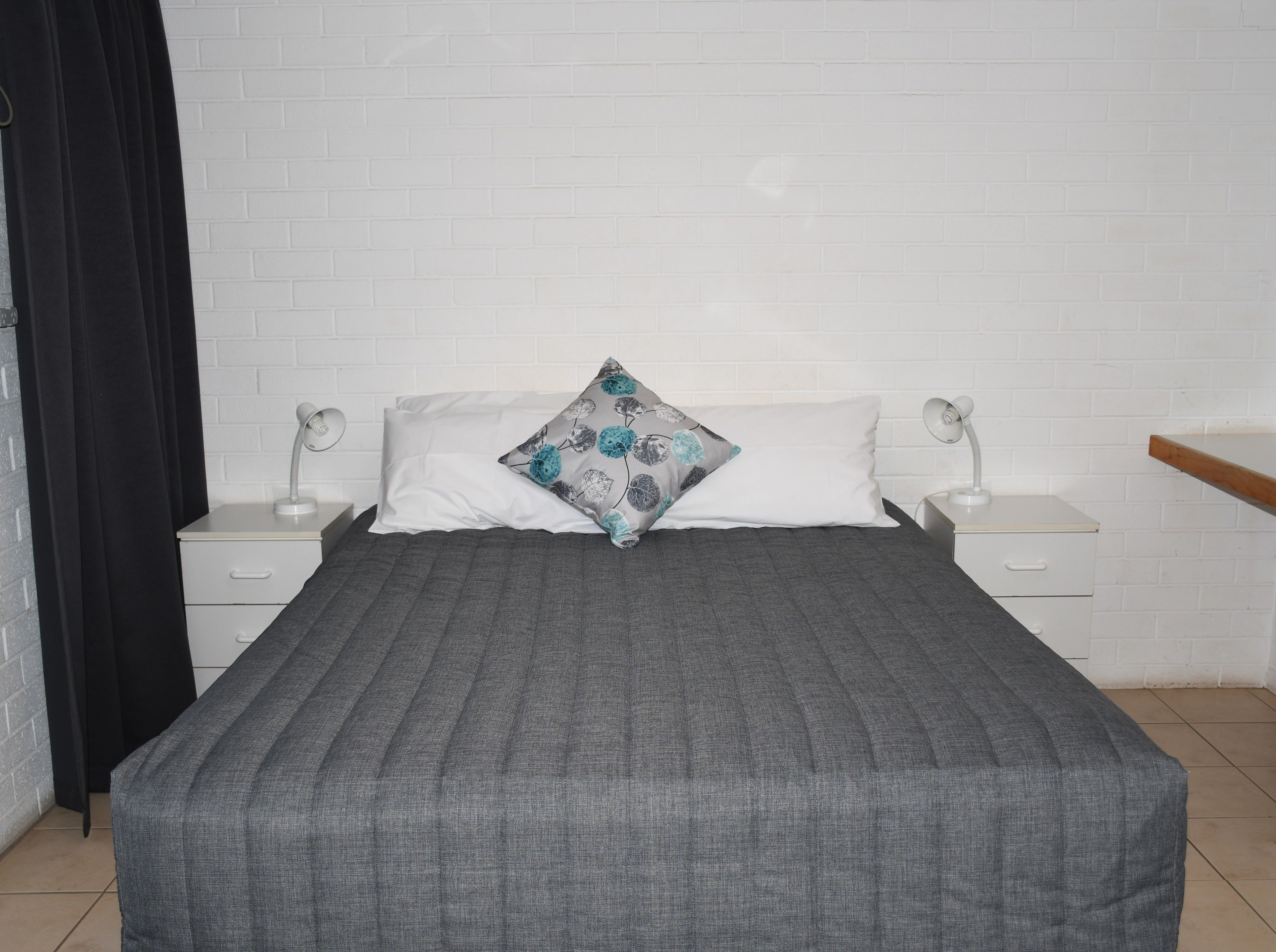 A bed with a grey quilt, two bedside tables with lamps, a white brick wall, and a black curtain.
