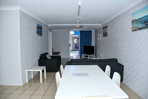 A modern living room with a white tiled floor, grey brick walls, a white dining table with chairs, a black sofa, and a flat-screen TV.