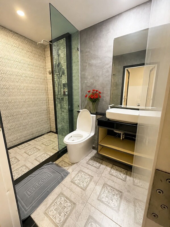 A modern bathroom with a glass shower enclosure, a white toilet, a sink with a mirror, and patterned tiles. - The Mantra Hotel (Karon)