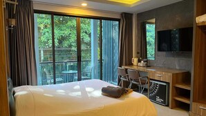 A hotel room with a bed, desk, chairs, a TV, and large windows with a view of greenery. - The Mantra Hotel (Karon)