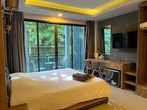 Une chambre d’hôtel équipée d’un lit, d’un bureau, de chaises, d’une télévision et de grandes fenêtres donnant sur un paysage verdoyant.