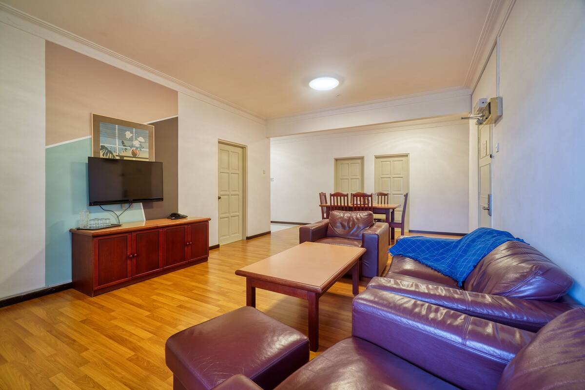 A living room with a brown leather sofa, a wooden coffee table, a TV on a wooden cabinet, and a blue blanket on the sofa.