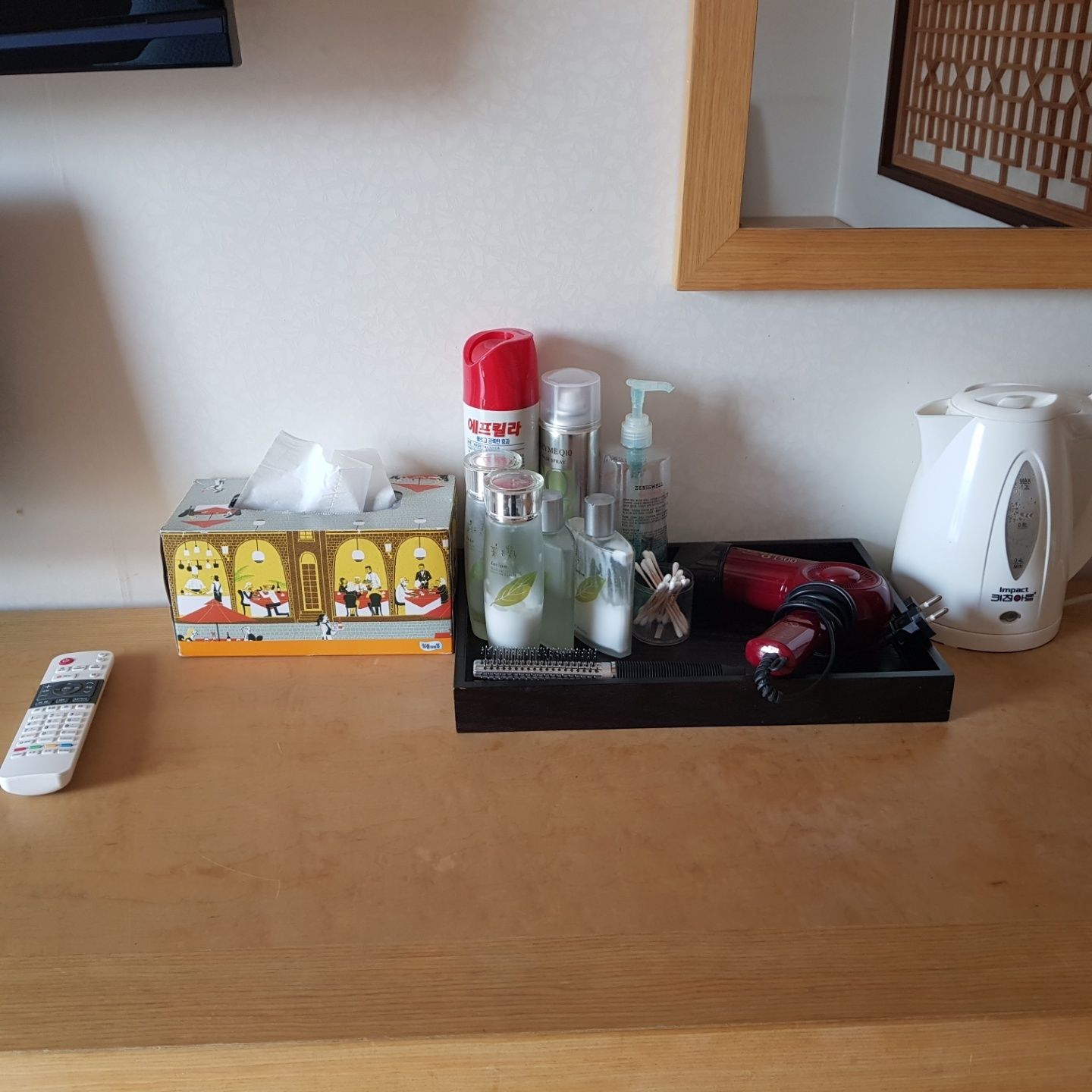 Una mesa con un control remoto, una caja de pañuelos, una bandeja con artículos de tocador y una tetera eléctrica blanca.