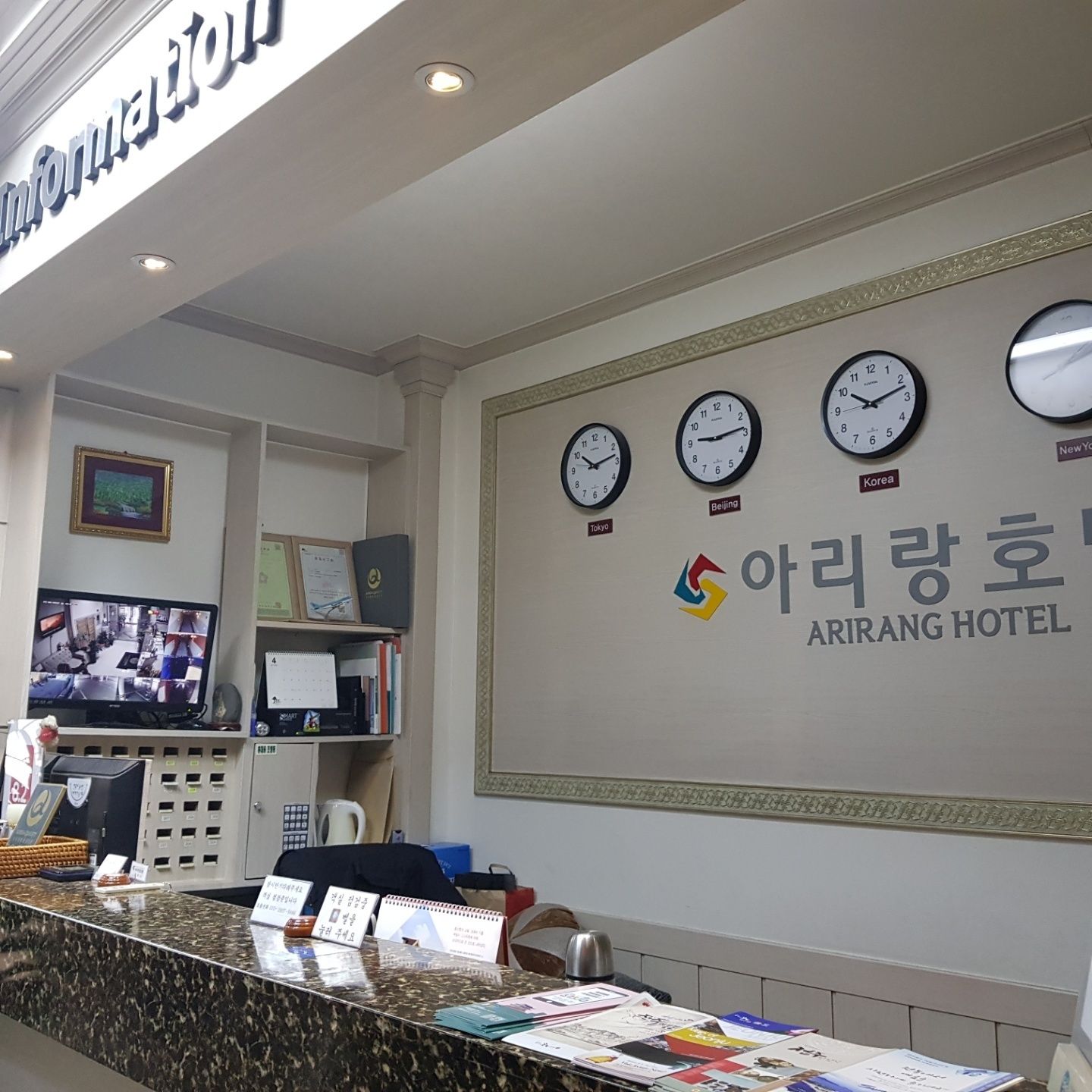 Zona de recepción de un hotel, con mostrador de recepción, un televisor mostrando imágenes de cámaras de seguridad y relojes que indican diferentes zonas horarias.