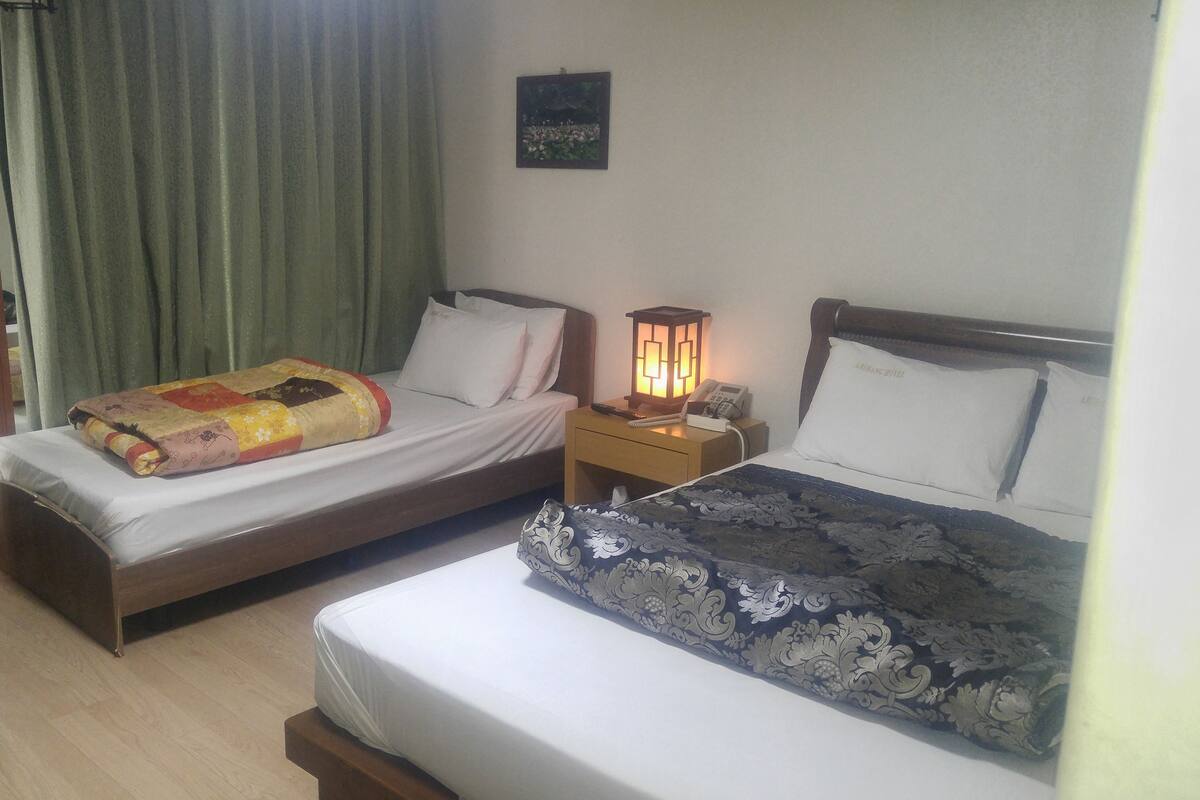 Habitación de hotel con dos camas individuales, una mesita de noche de madera con una lámpara, una silla y una pequeña mesa.