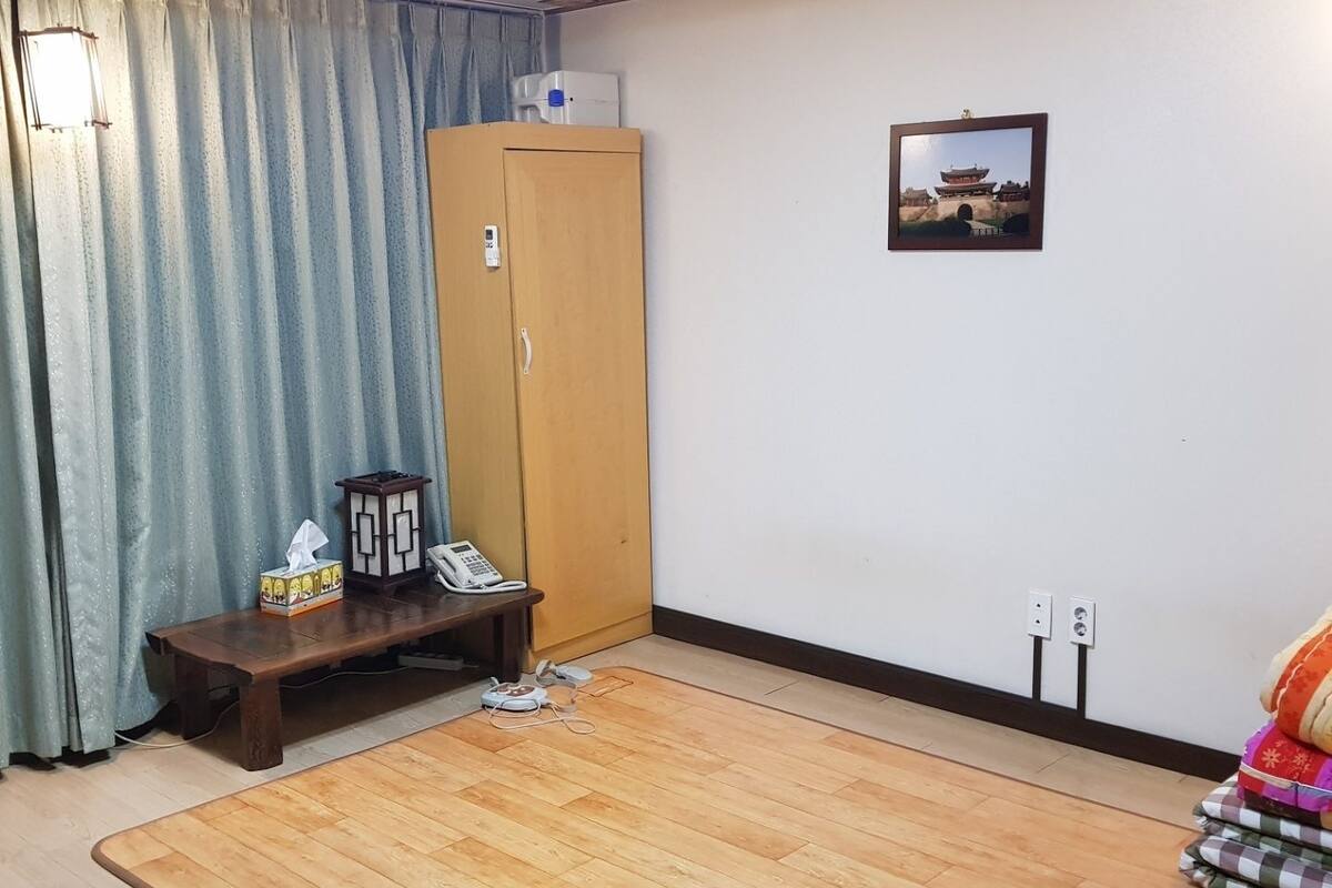 Una habitación con piso de madera, un armario, una pequeña mesa con teléfono y caja de pañuelos, un cuadro en la pared y una cortina.