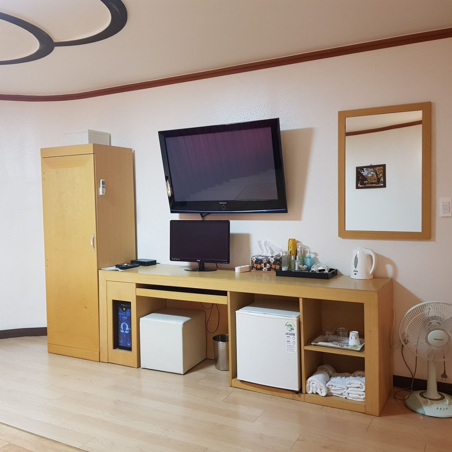 A compact room with a wooden desk, a television, a small refrigerator, a cabinet, a fan, and a framed picture on the wall.