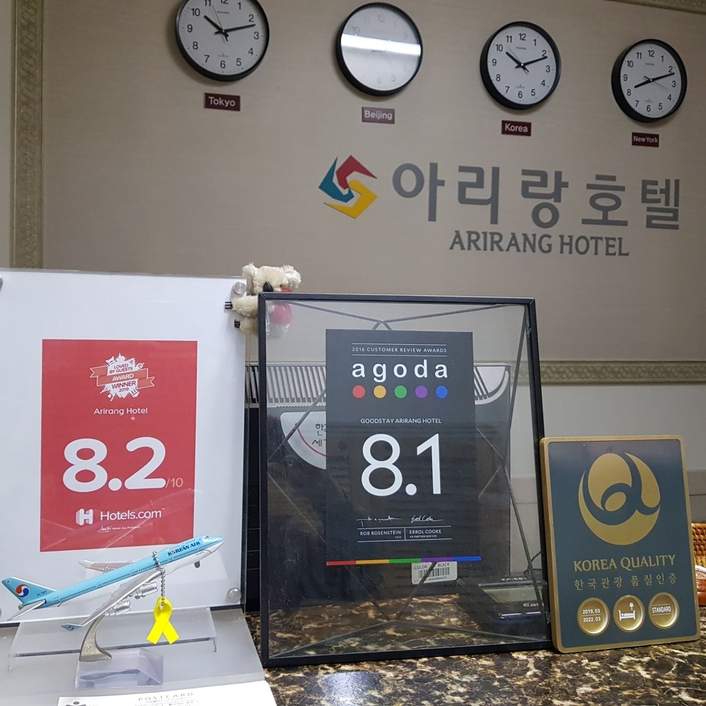 A hotel reception area with clocks displaying different time zones, a sign for Arirang Hotel, and awards for customer service.