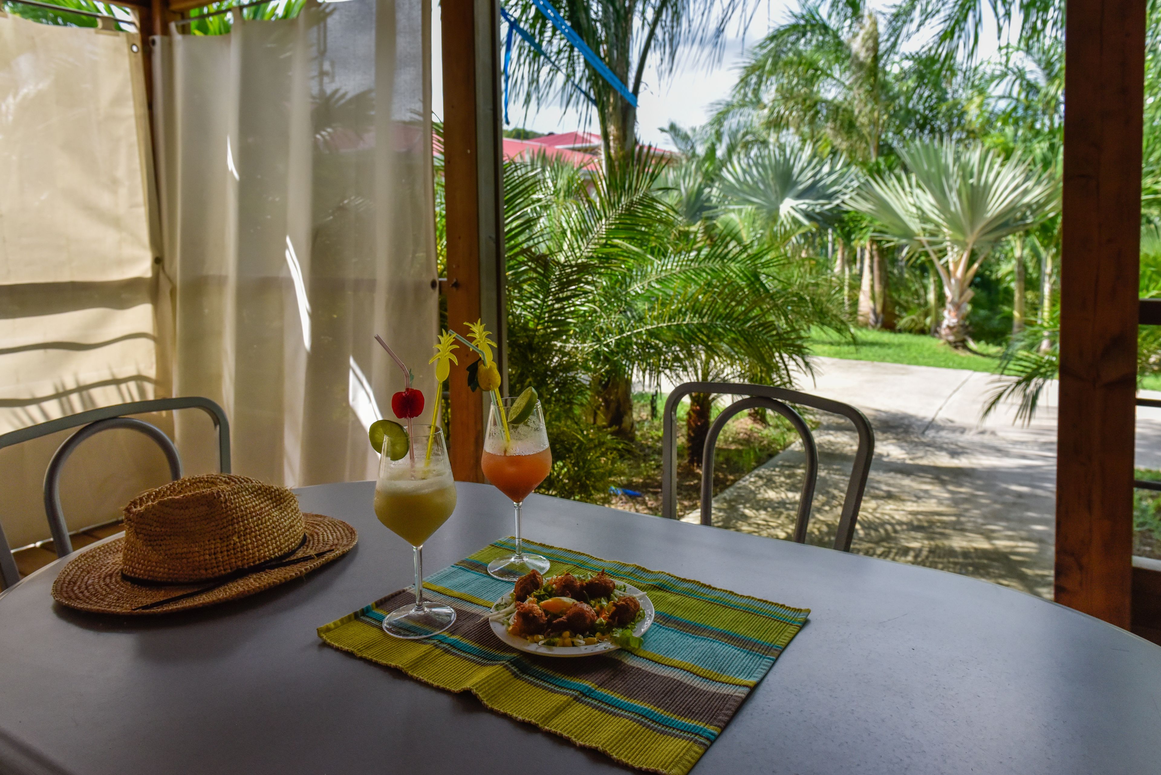 A table with a straw hat, two drinks, and a plate of food, set against a backdrop of lush greenery and a view of the road.