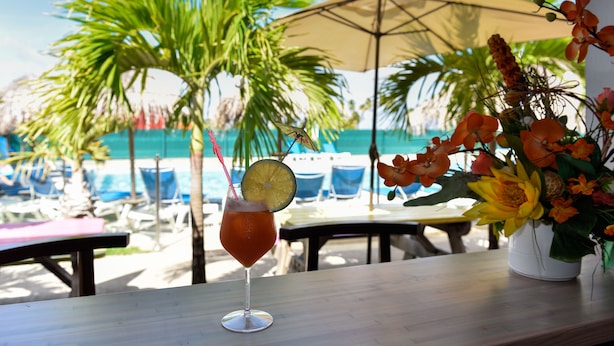 A tropical setting with a drink on a table, palm trees, and a pool in the background.