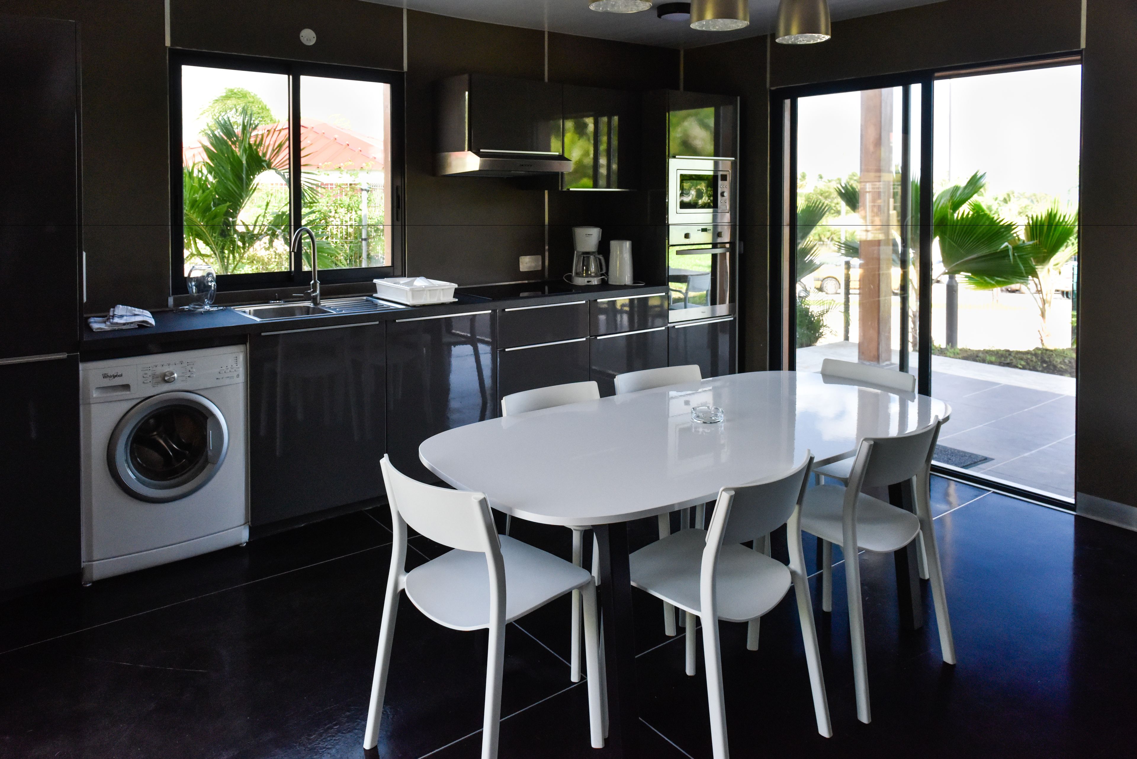 Una cocina moderna con lavadora integrada, un comedor con sillas blancas y ventanas amplias que ofrecen vista a la vegetación exterior.