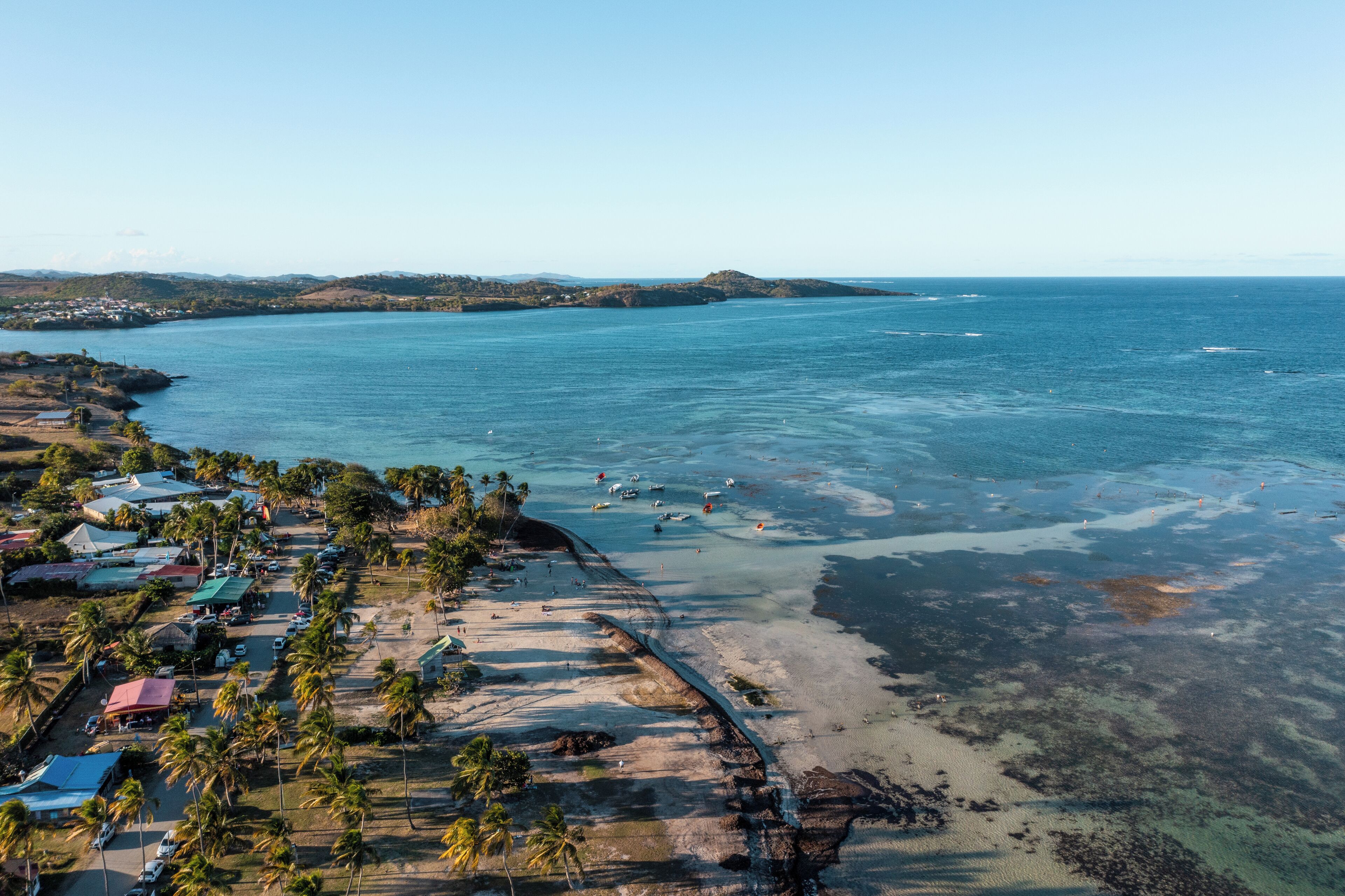 An aerial view of a coastal area with a beach, clear blue water, and a small settlement with buildings and palm trees.