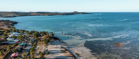 An aerial view of a coastal area with a beach, clear blue water, and a small settlement with buildings and palm trees.