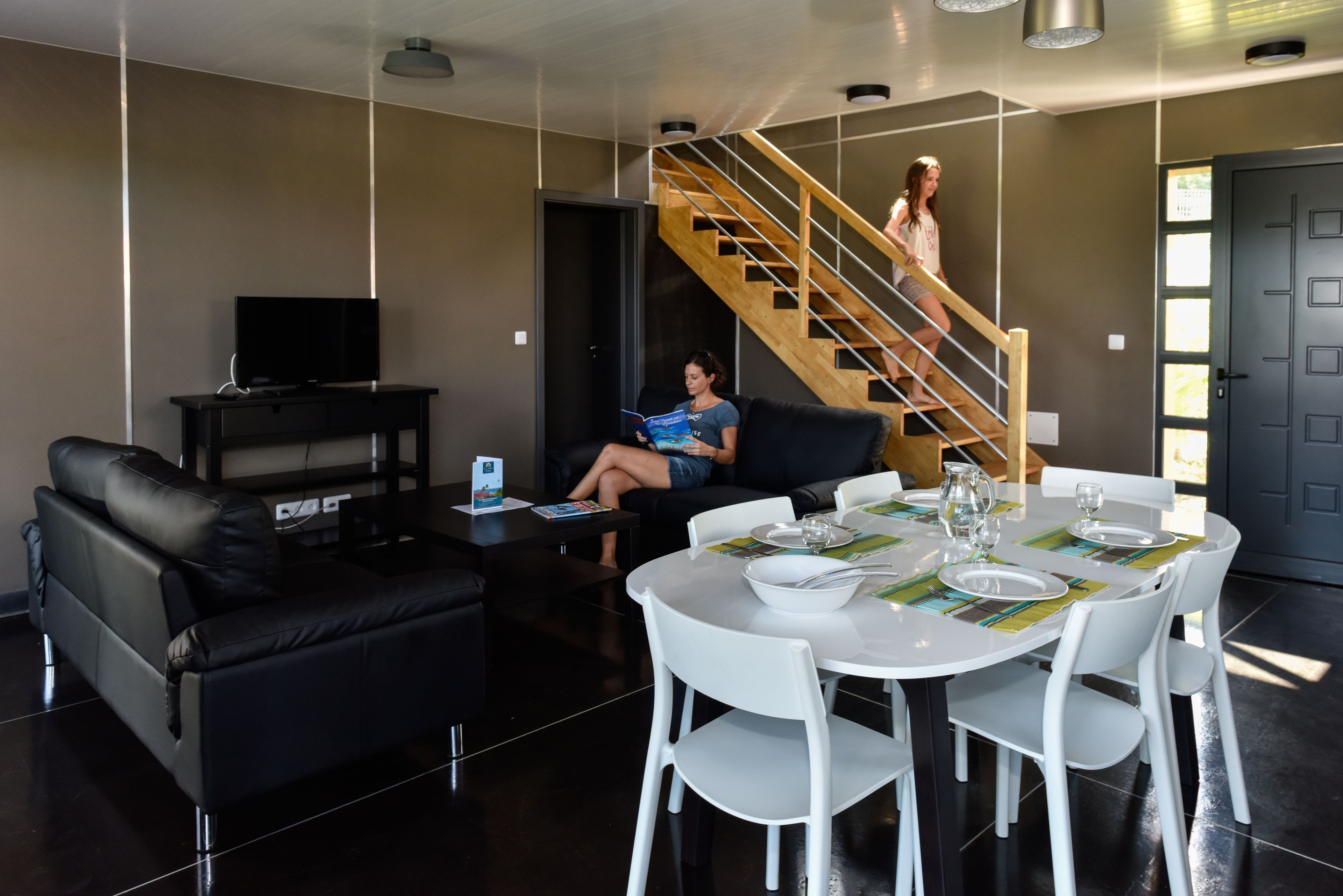 A modern living room with a black leather sofa, a white dining table set for four, and a staircase leading to an upper level.
