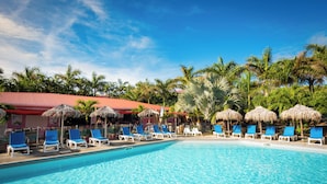 A swimming pool with blue water, surrounded by lounge chairs and thatched umbrellas, with palm trees and a red-roofed building in the background.