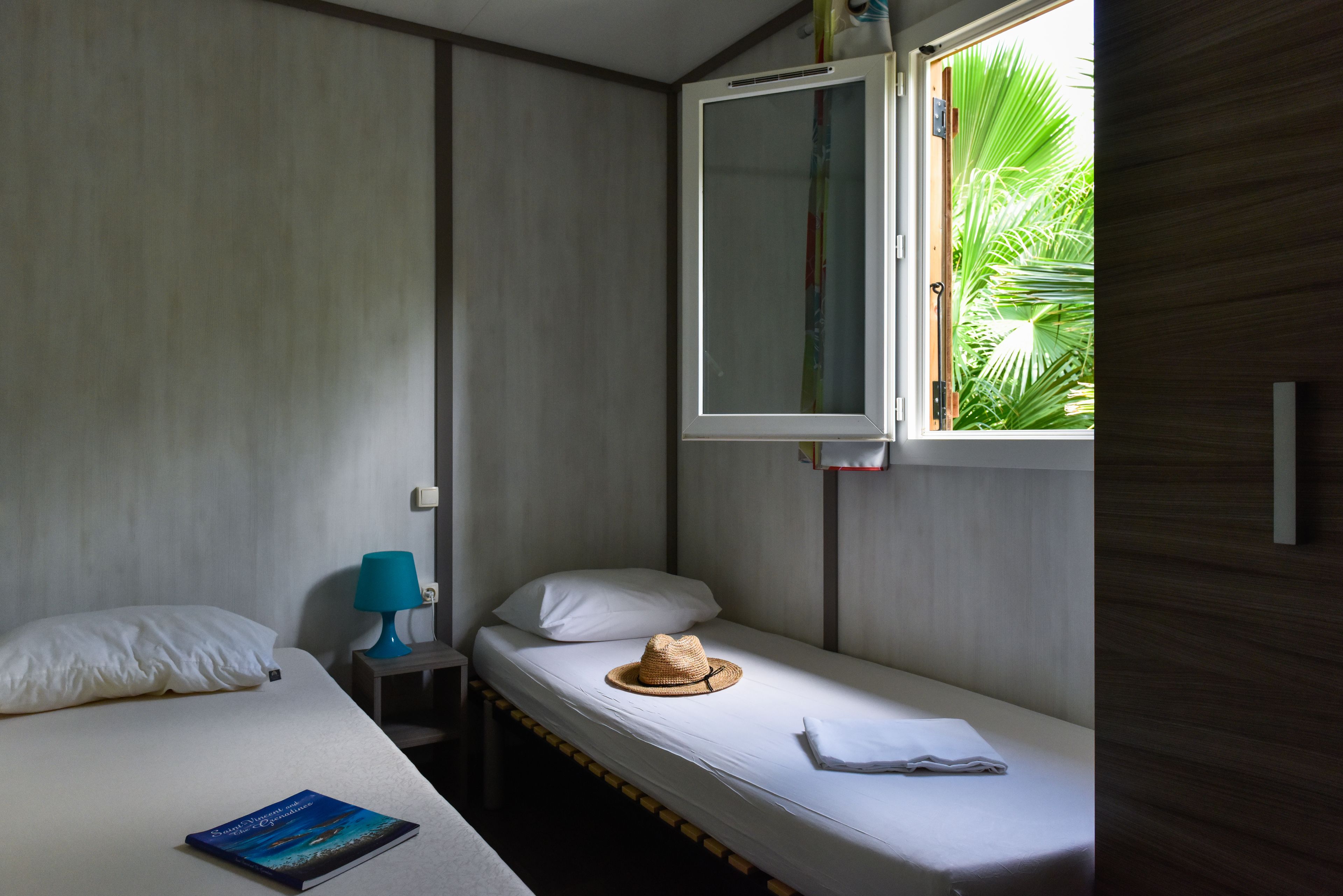 A single-bed room with a window, a bedside table with a lamp, and a hat on the bed.