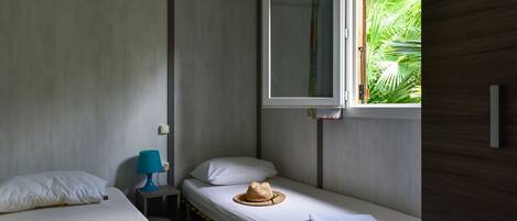 A single-bed room with a window, a bedside table with a lamp, and a hat on the bed.