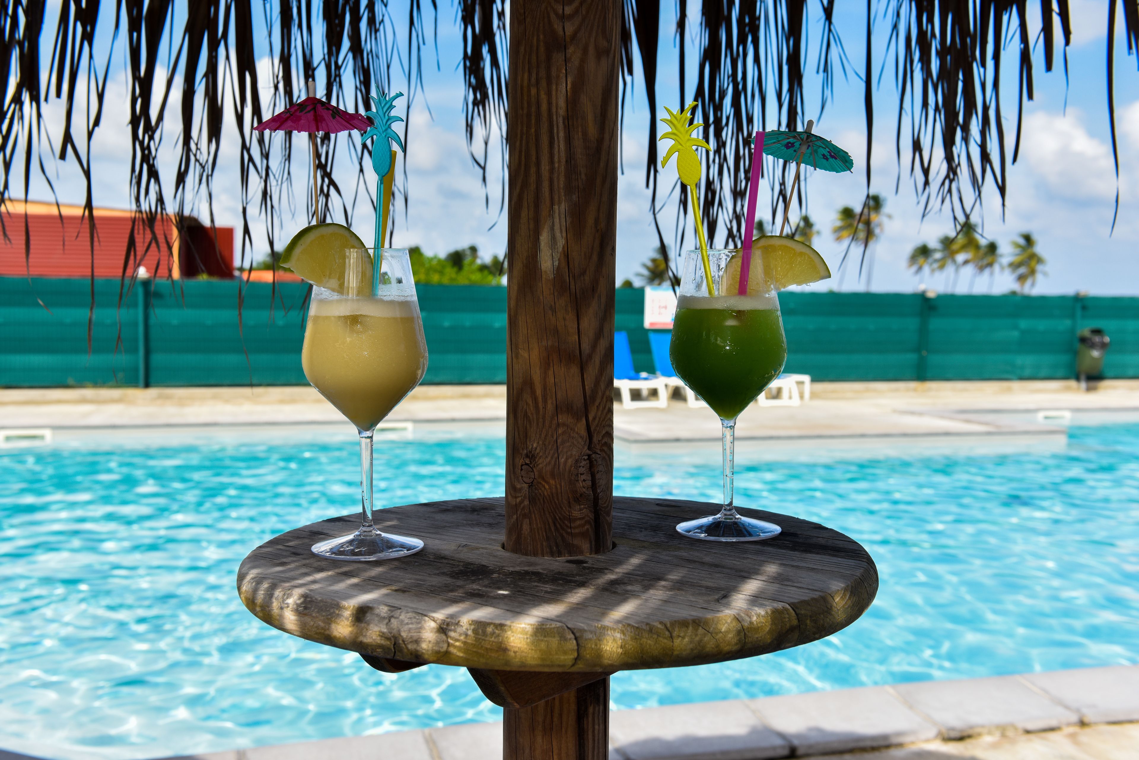 Dos cócteles sobre una mesa de madera junto a una piscina, con una palmera al fondo.