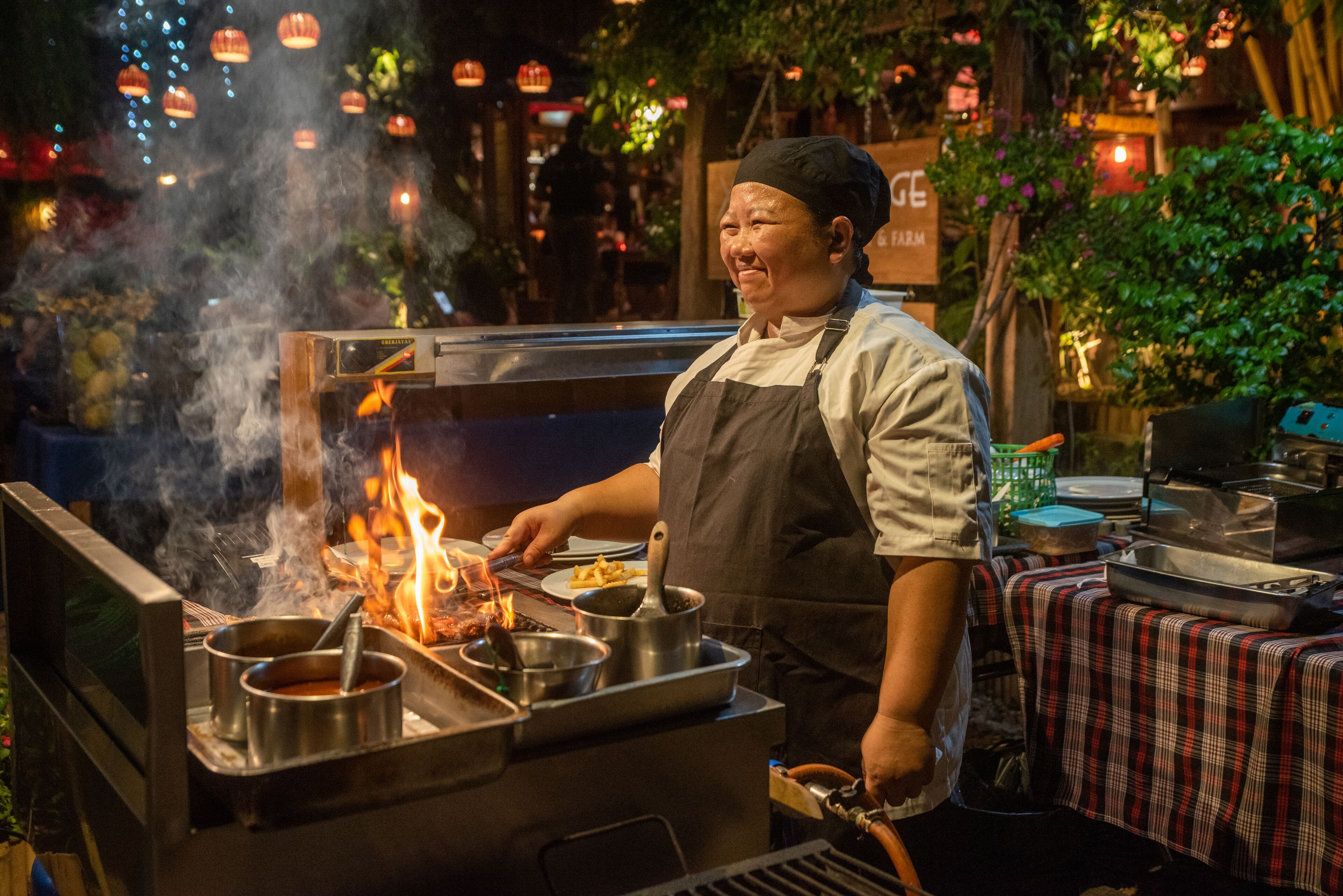 A chef cooking outdoors with a portable stove, surrounded by pots and pans, and a table with a checkered tablecloth.
