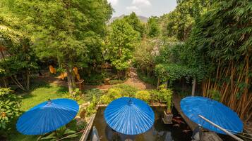 Un jardin avec un étang, entouré d’une végétation luxuriante et de bambous. On y trouve trois parasols bleus qui offrent de l’ombre.