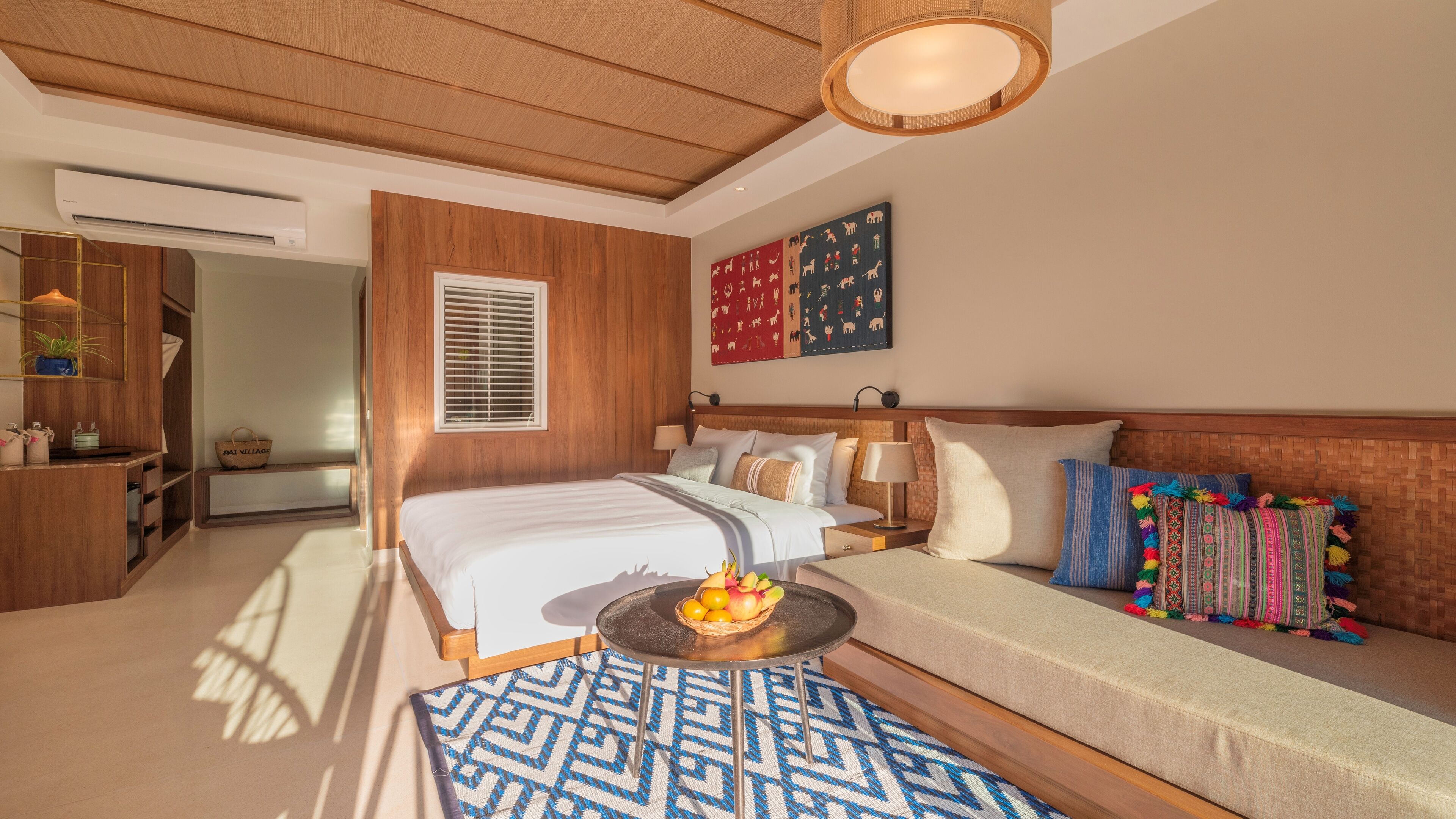 A hotel room with a wooden ceiling, a bed with white bedding, a sofa with colorful pillows, a small round table with fruit, and a patterned rug.
