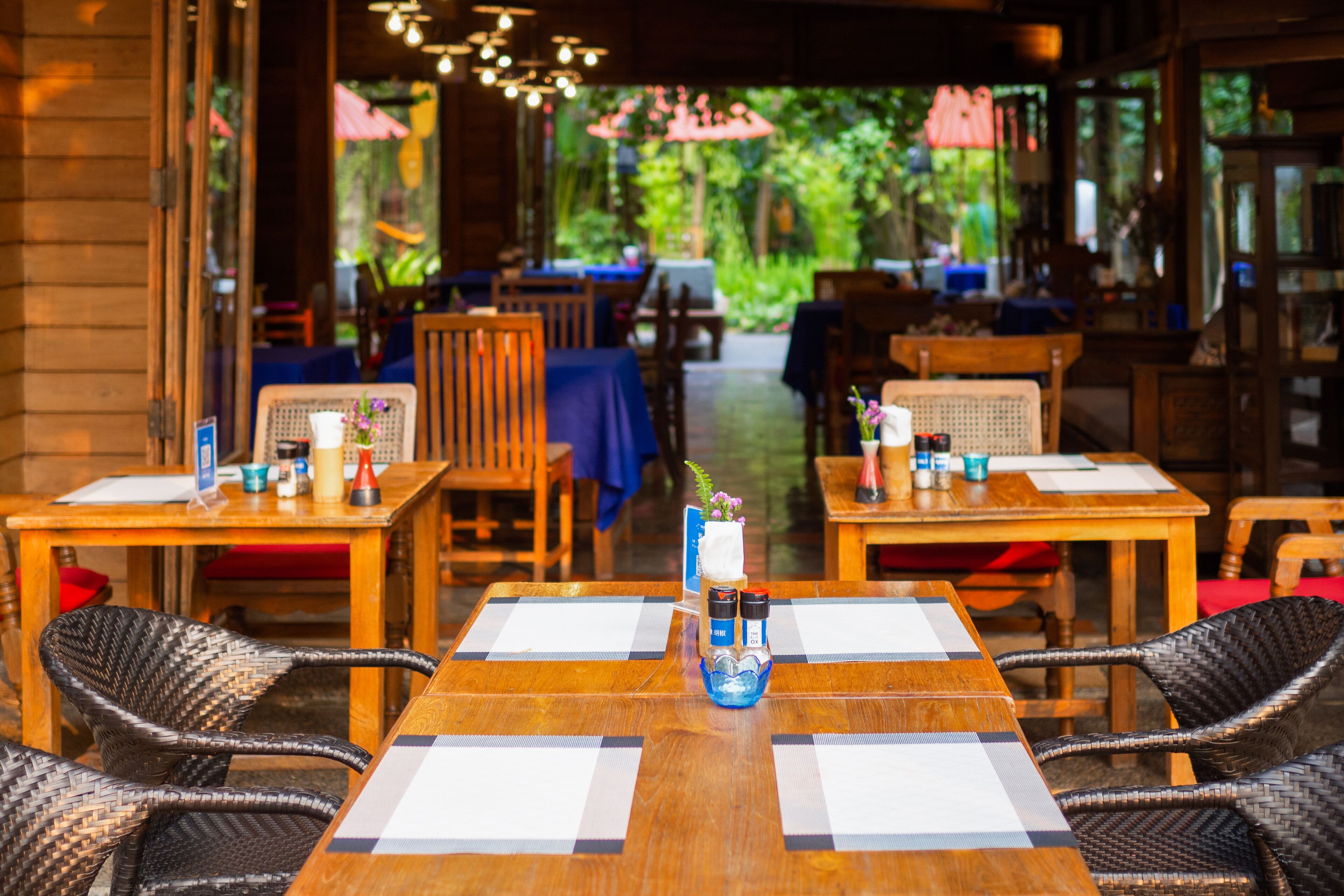 An outdoor dining area with wooden tables and wicker chairs, featuring a blue vase with flowers and condiments.