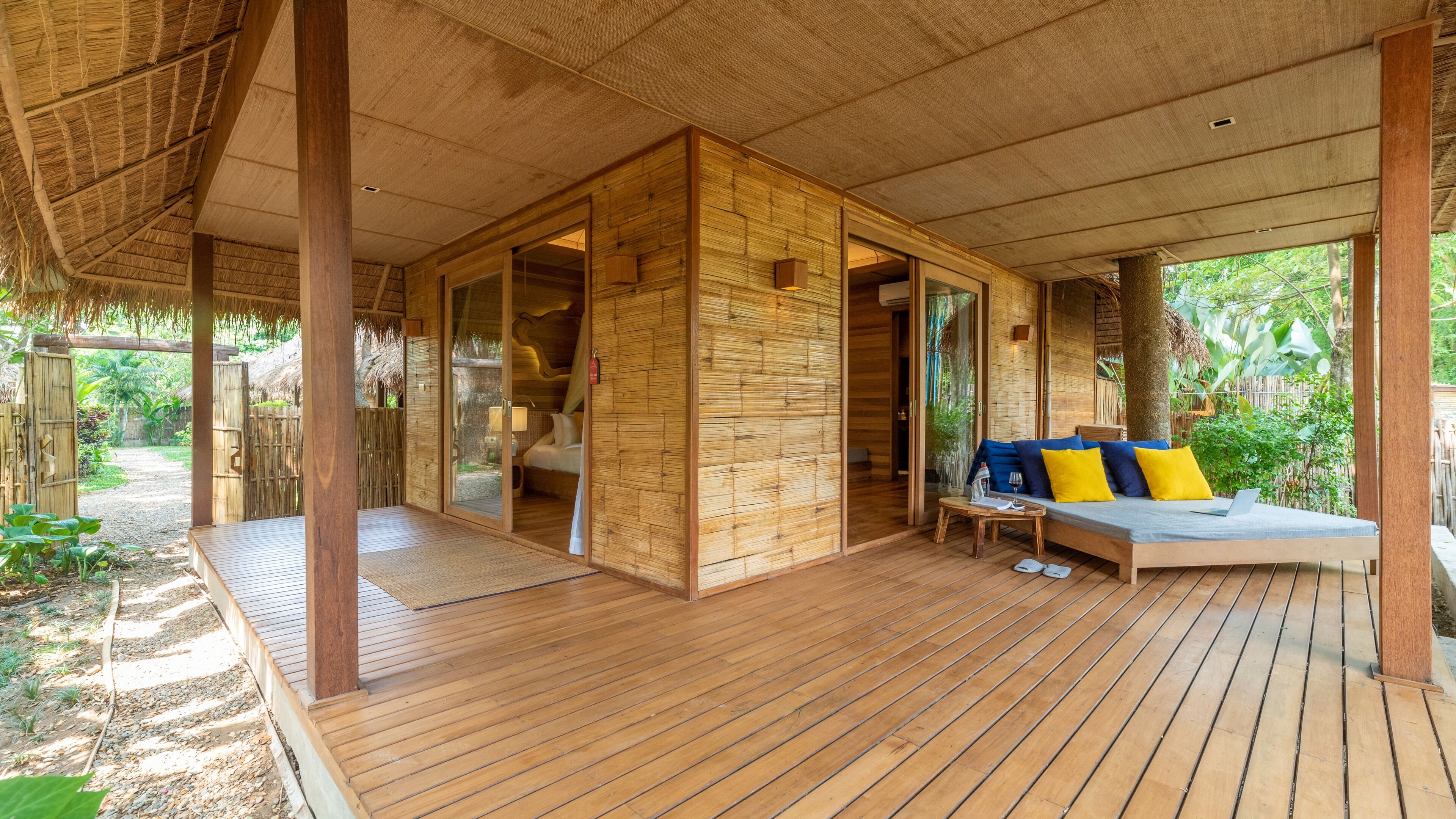 Une cabane en bois avec un toit de chaume, une terrasse en bois et un canapé avec des coussins.