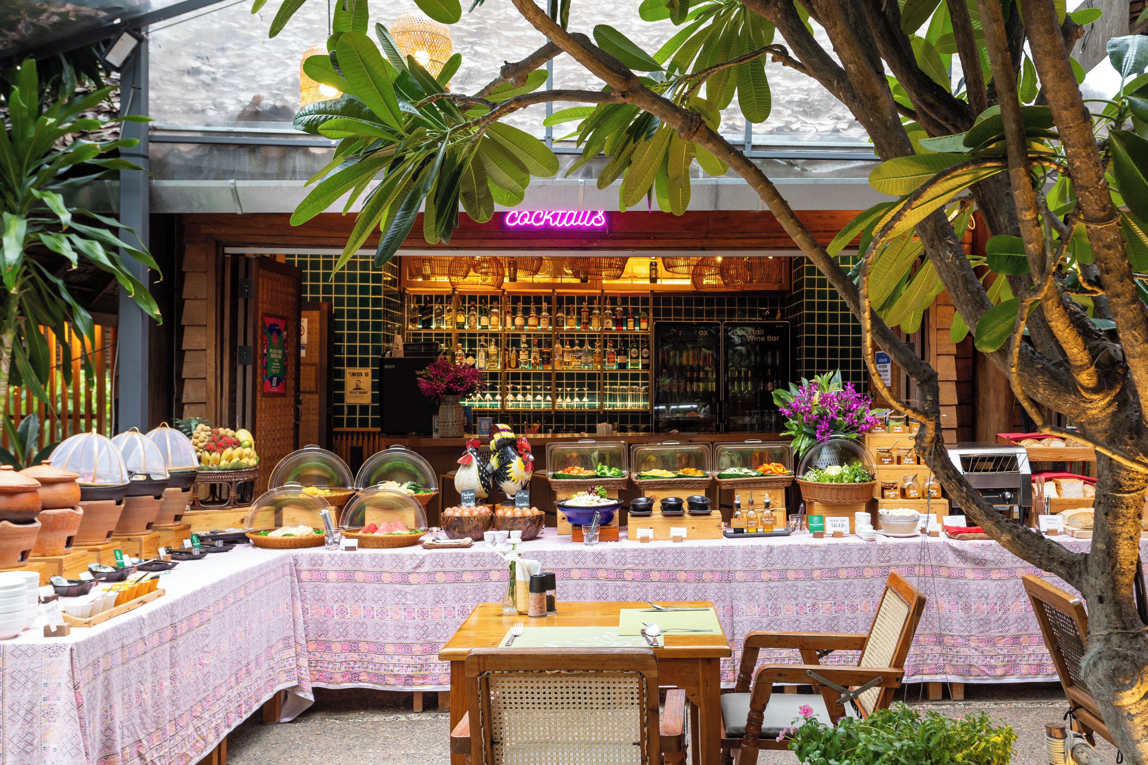 An outdoor dining area with a buffet setup, a bar with a neon sign, and tropical plants.