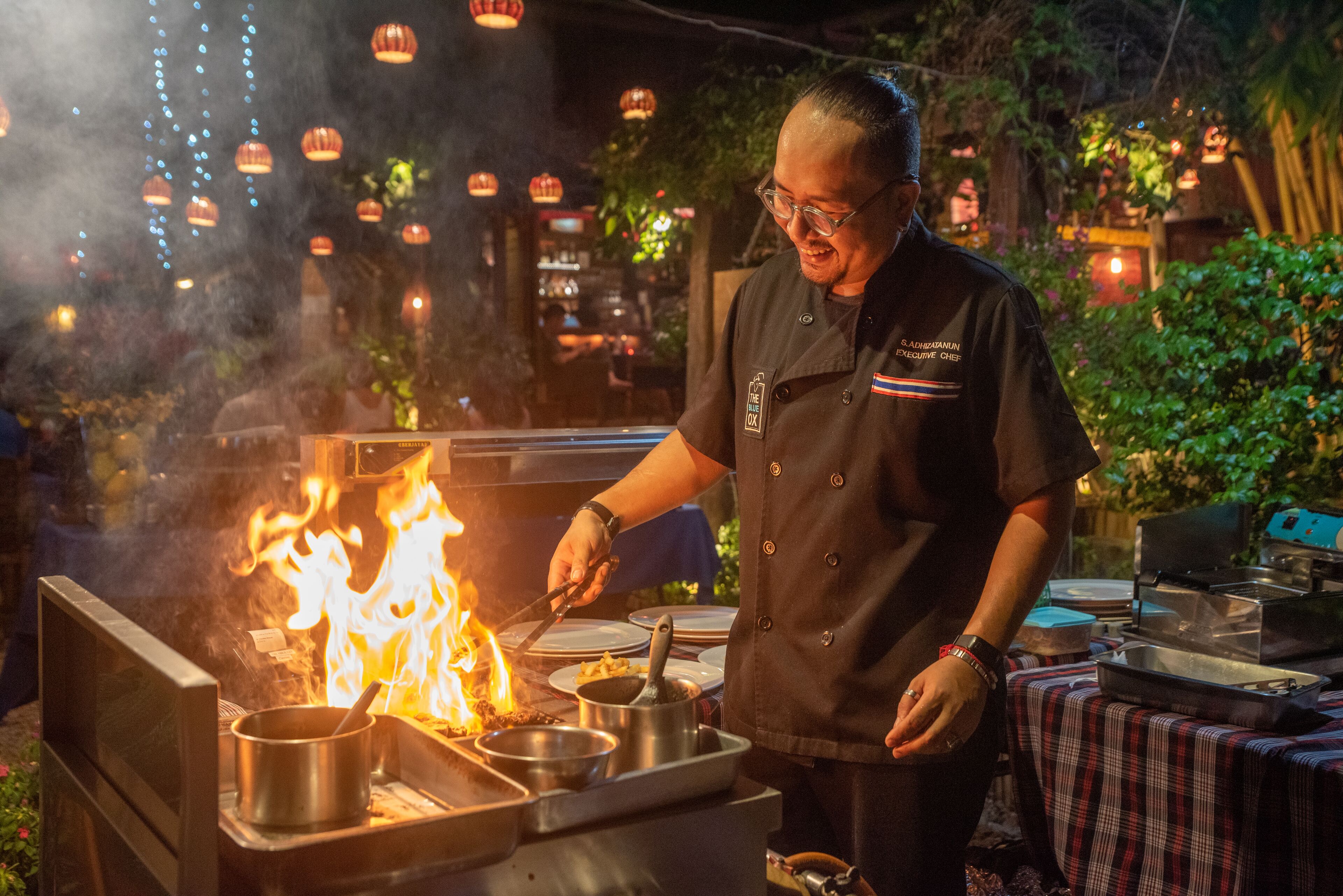 A chef cooking outdoors with a portable stove and pots, surrounded by hanging lanterns and greenery.