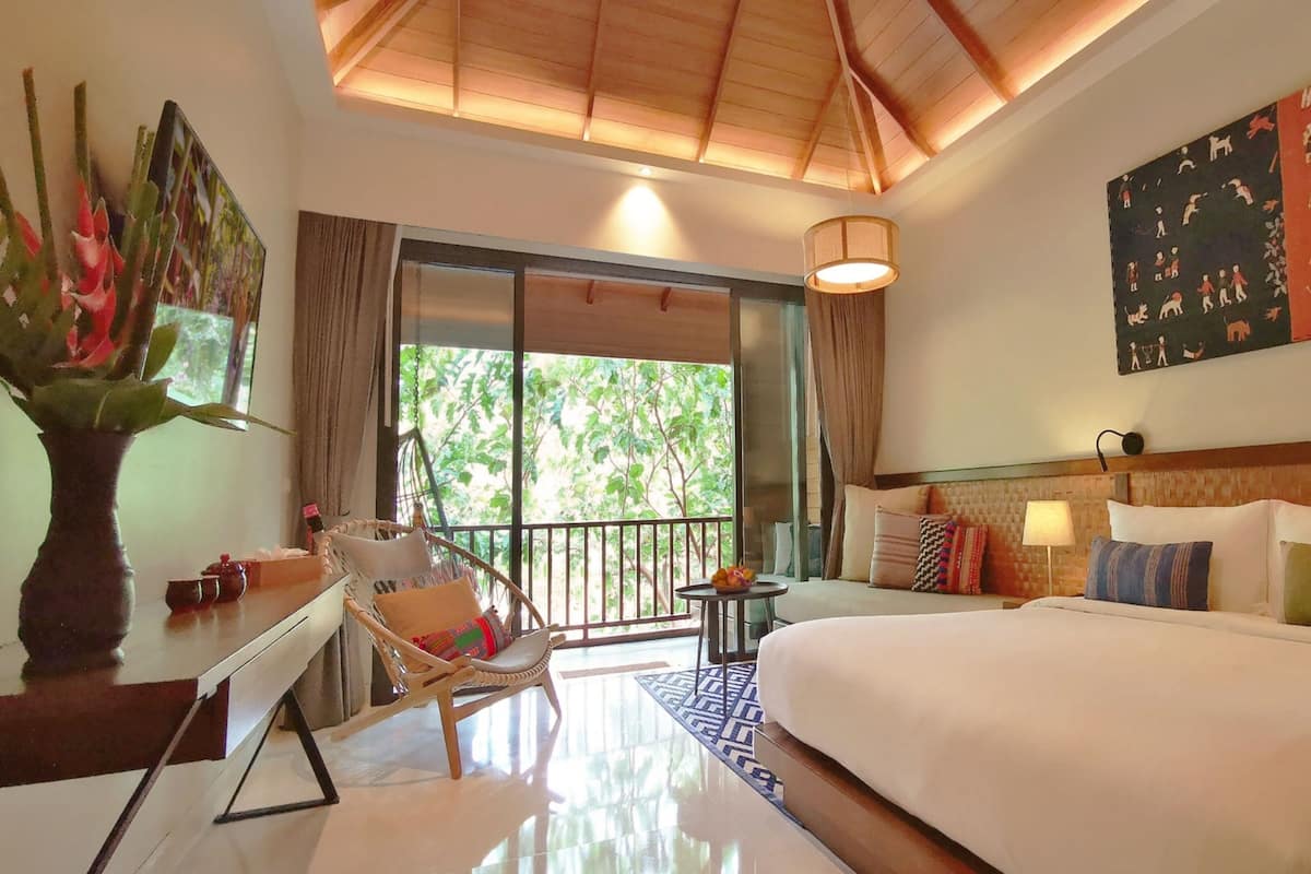 A hotel room with a bed, a desk, a chair, a small table with fruit, and a large window with a view of greenery.