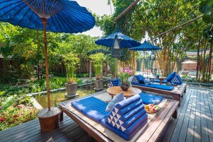 A wooden deck with blue cushioned seating and umbrellas, surrounded by lush greenery and a small pond.
