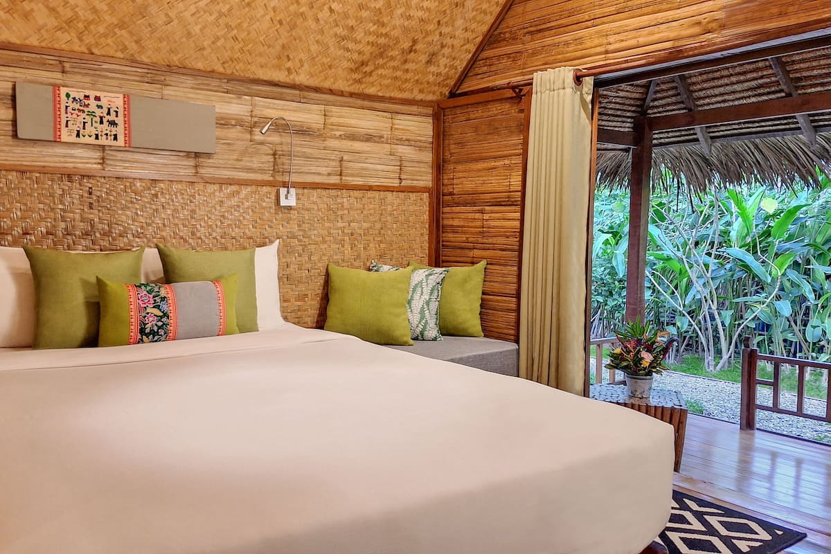 A room with a large bed, green and white pillows, a wooden ceiling, and a view of greenery outside.