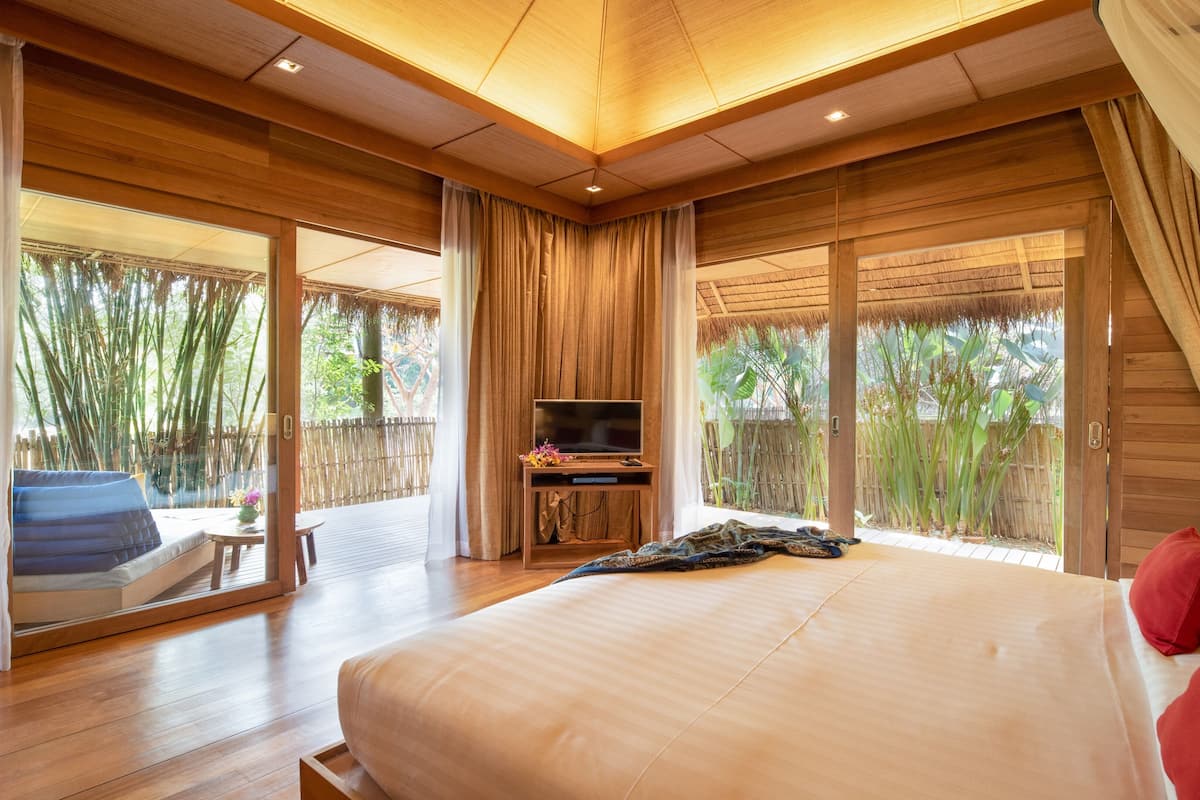 A spacious bedroom with a large bed, a TV on a wooden stand, and a balcony with bamboo views.
