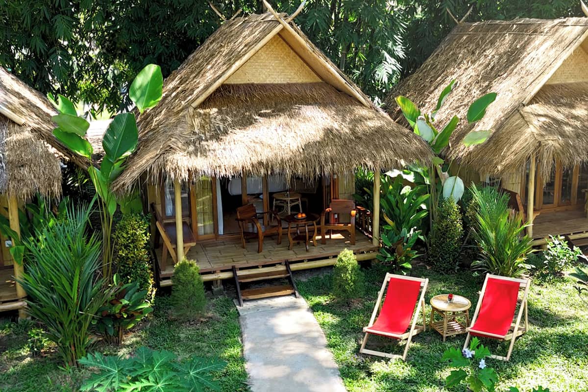 A thatched-roof bungalow with a wooden deck, outdoor seating, and lush greenery.
