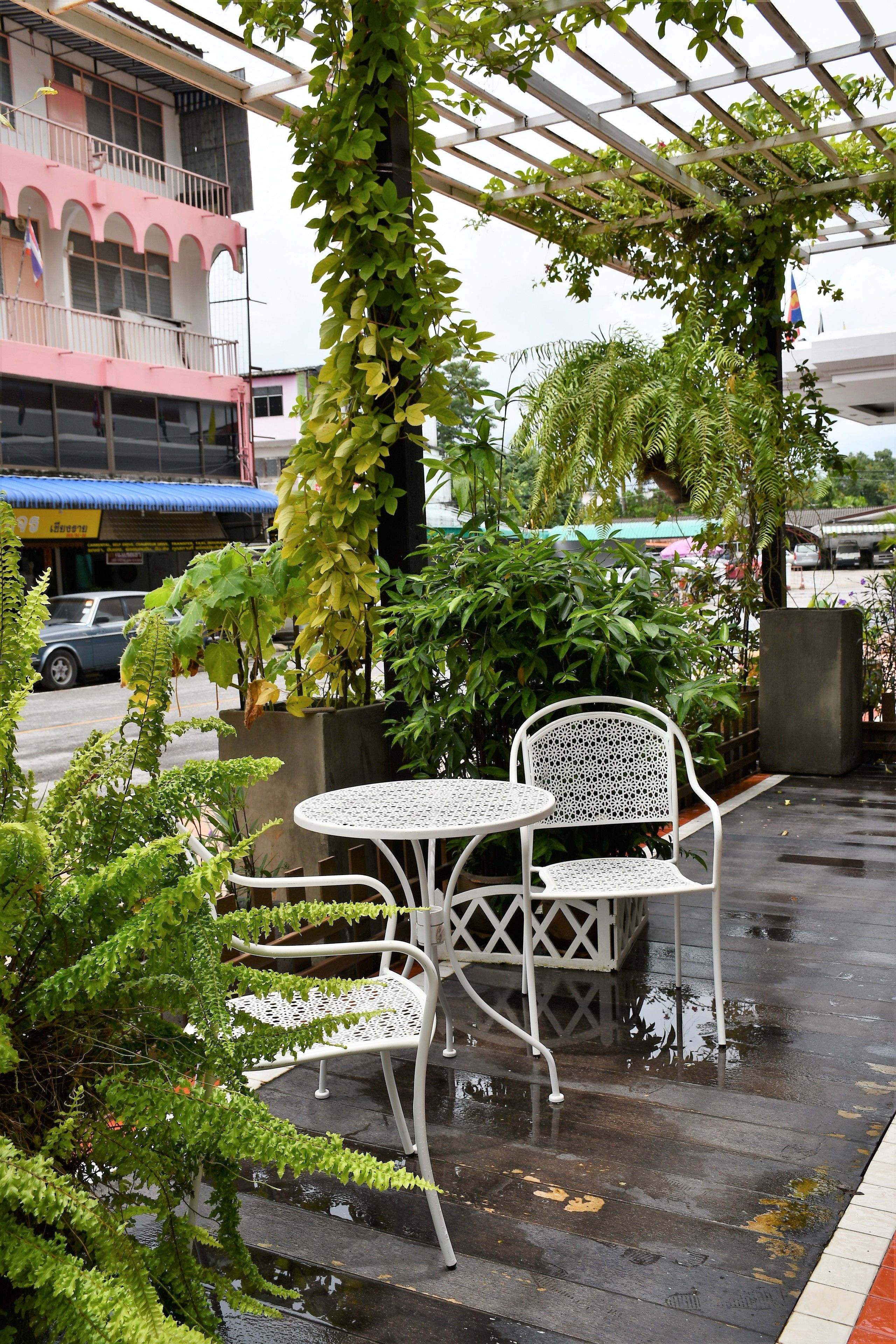 A covered outdoor seating area with a table and chairs, surrounded by lush greenery and a wet pavement.