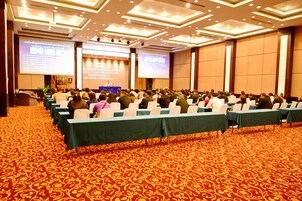 Un salón de conferencias con filas de mesas y sillas, piso alfombrado y una pantalla grande al frente.