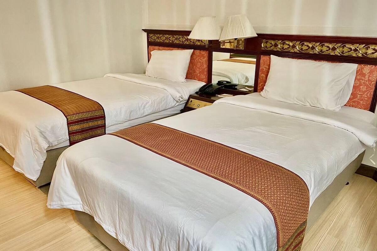 A hotel room with two beds, each with white linens and a patterned runner. There is a wooden headboard with a decorative design and two bedside lamps.