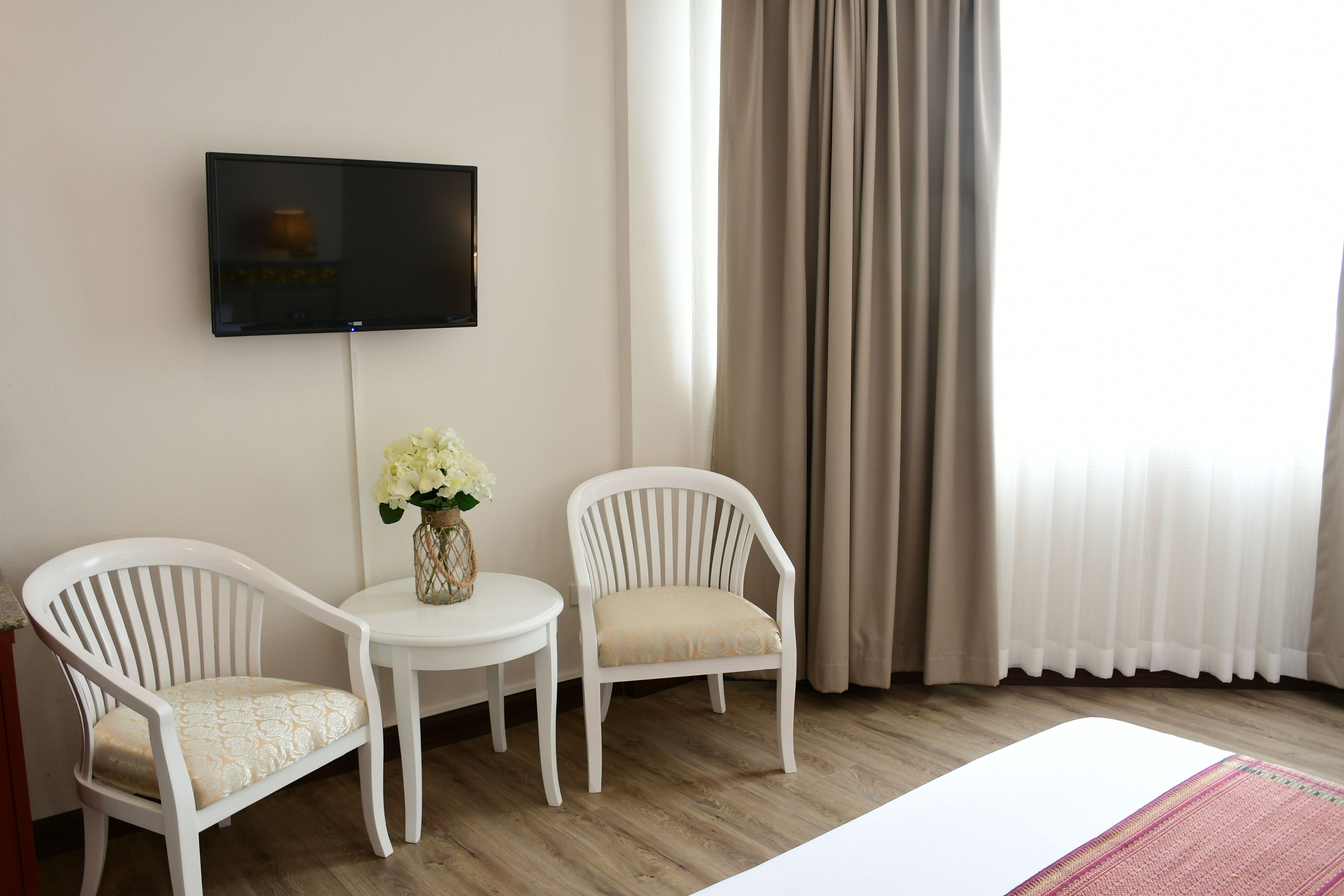 A hotel room with a flat-screen TV mounted on the wall, two white chairs with patterned cushions, a small round table with a vase of flowers, and a large window with beige curtains.