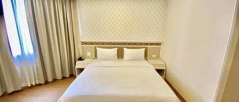 A hotel room with a large bed, two bedside tables, a nightstand, and a window with curtains.