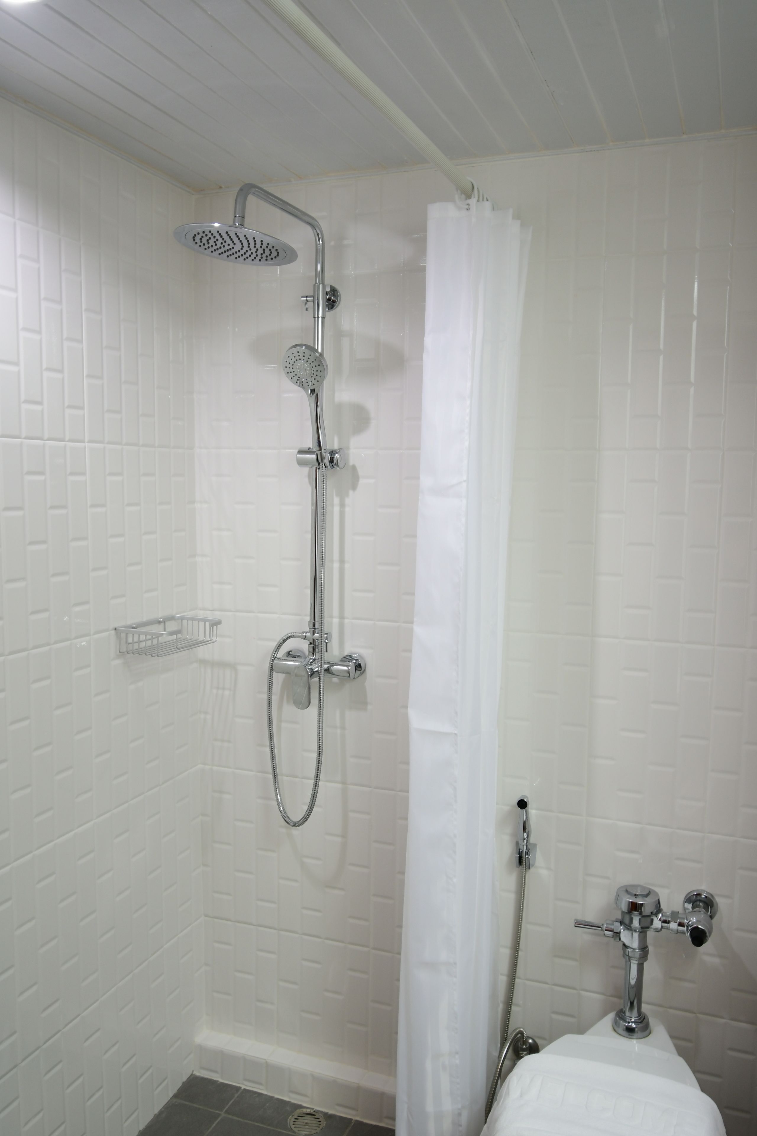 Une douche équipée d’un pommeau de douche à main et d’un robinet mural, d’un rideau de douche blanc et d’une petite étagère.