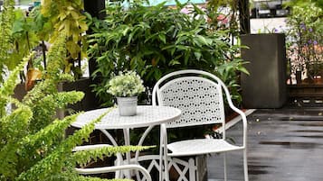A white metal table and chair set under a green vine, surrounded by lush plants and a wet floor.