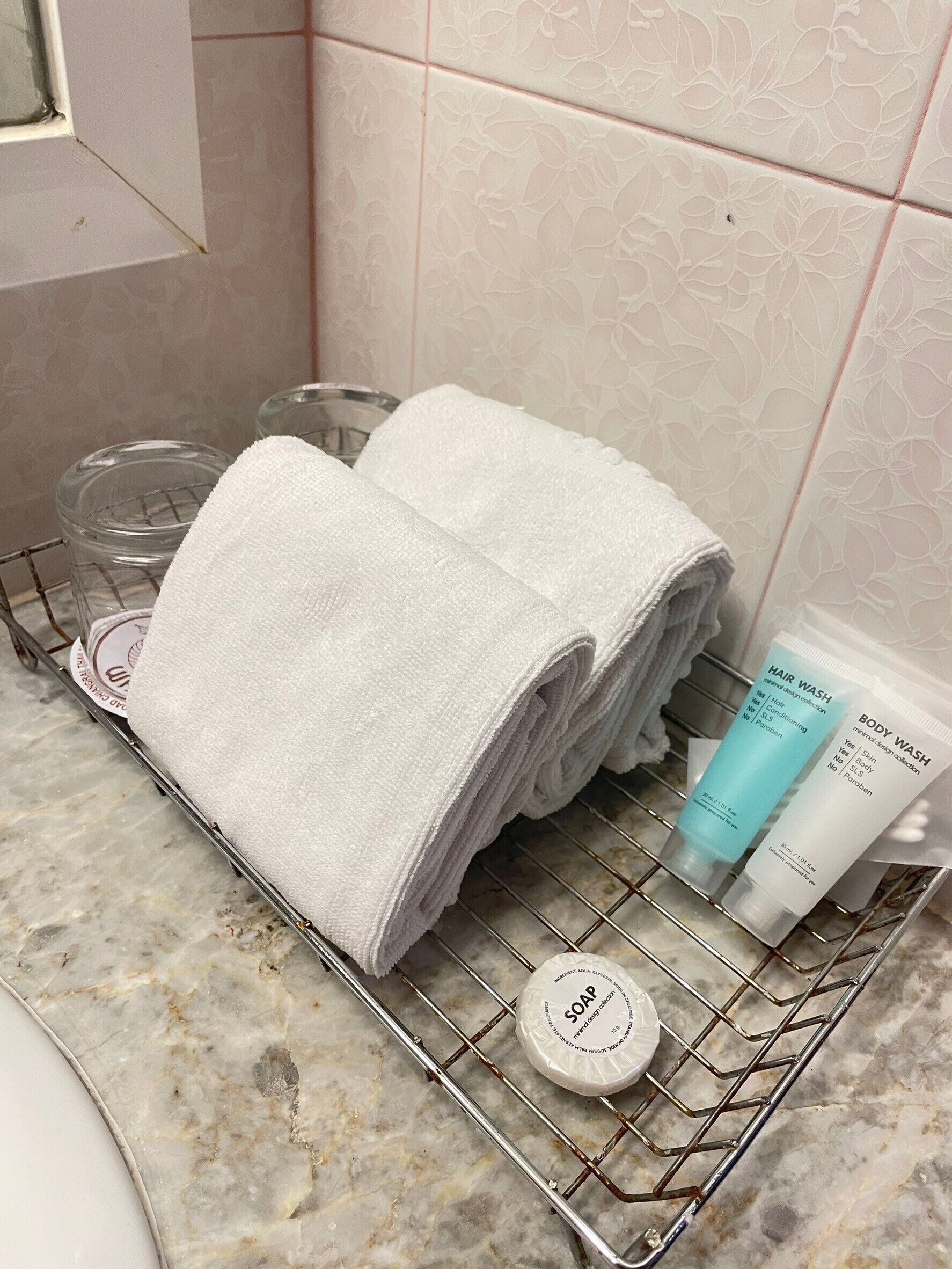 Un baño con encimera de mármol, un perchero con toallas dobladas, un recipiente de vidrio, una botella de gel de ducha, una botella de champú y un jabonera.